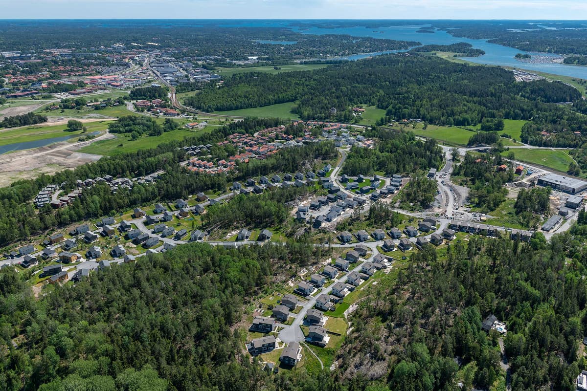 Vy från ovan över soligt villaområde med slingrande gator, allt omgivet av stora skogspartier.