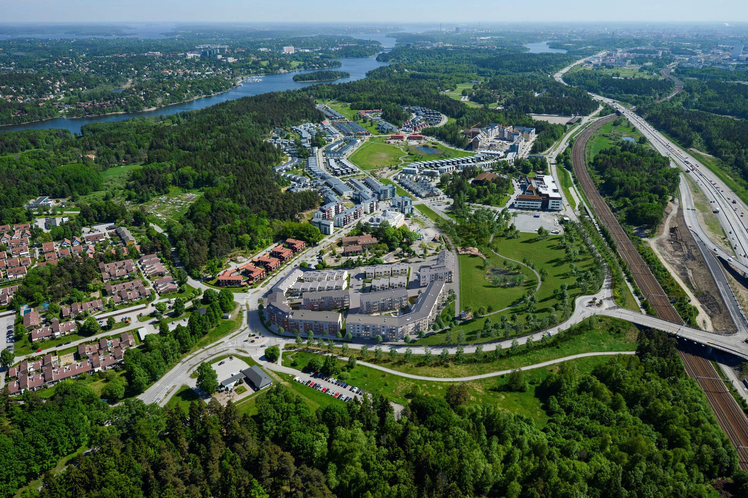 Flygvy över grönt och lummigt bostadsområde omgivet av skog och nära till glittrande vatten.