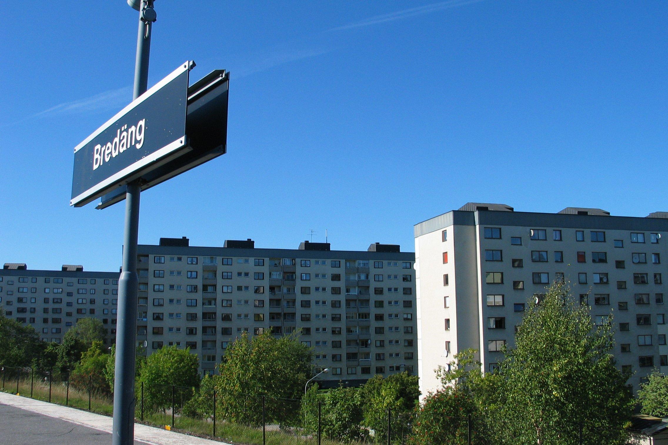 Flerbostadshus mot blå himmel och t-baneperrong med skylt där det står Bredäng.