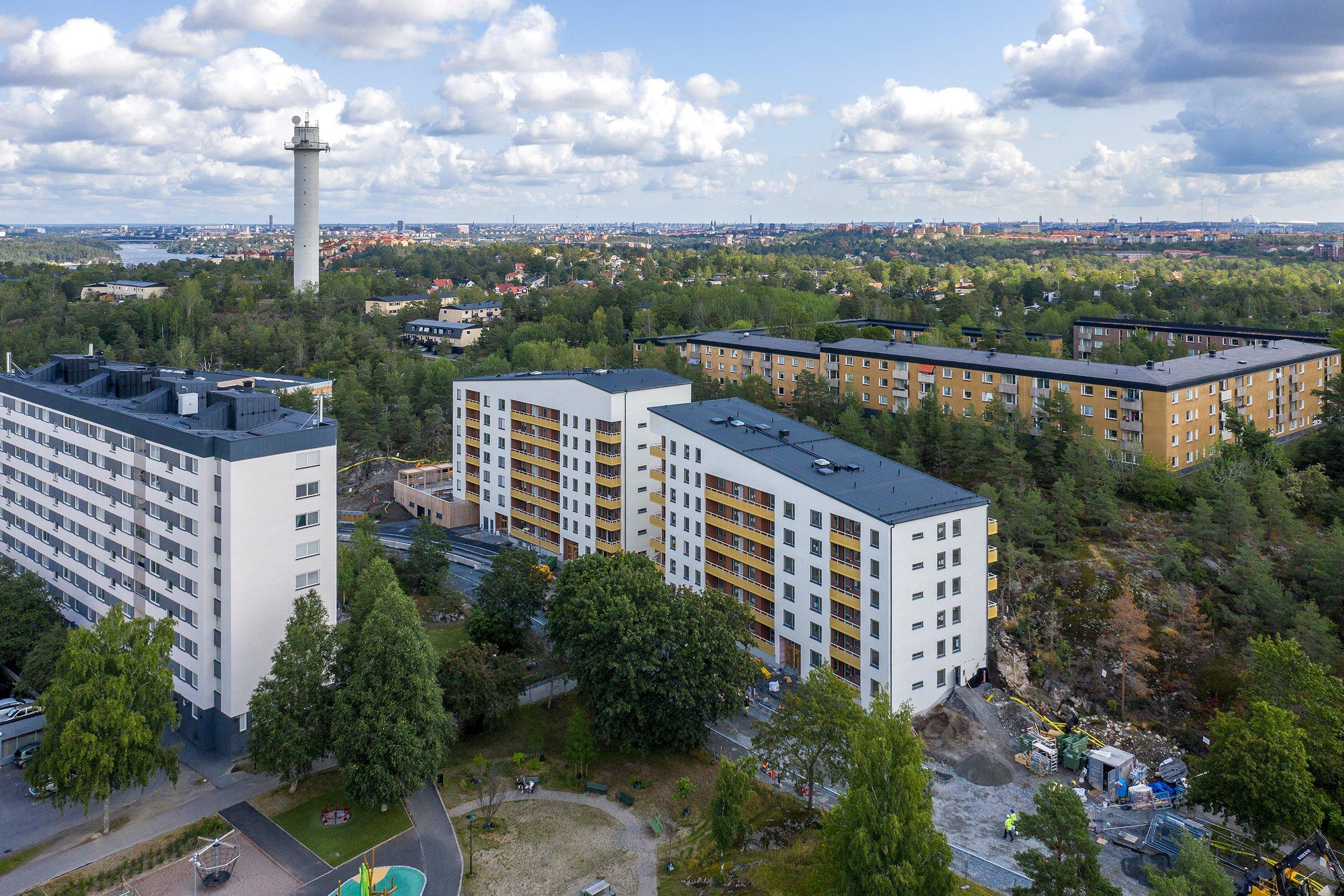 Vy från ovan över bostadsområde omgivet av mycket grönska. I fokus ligger några vita flerbostadshus med gula balkonger.