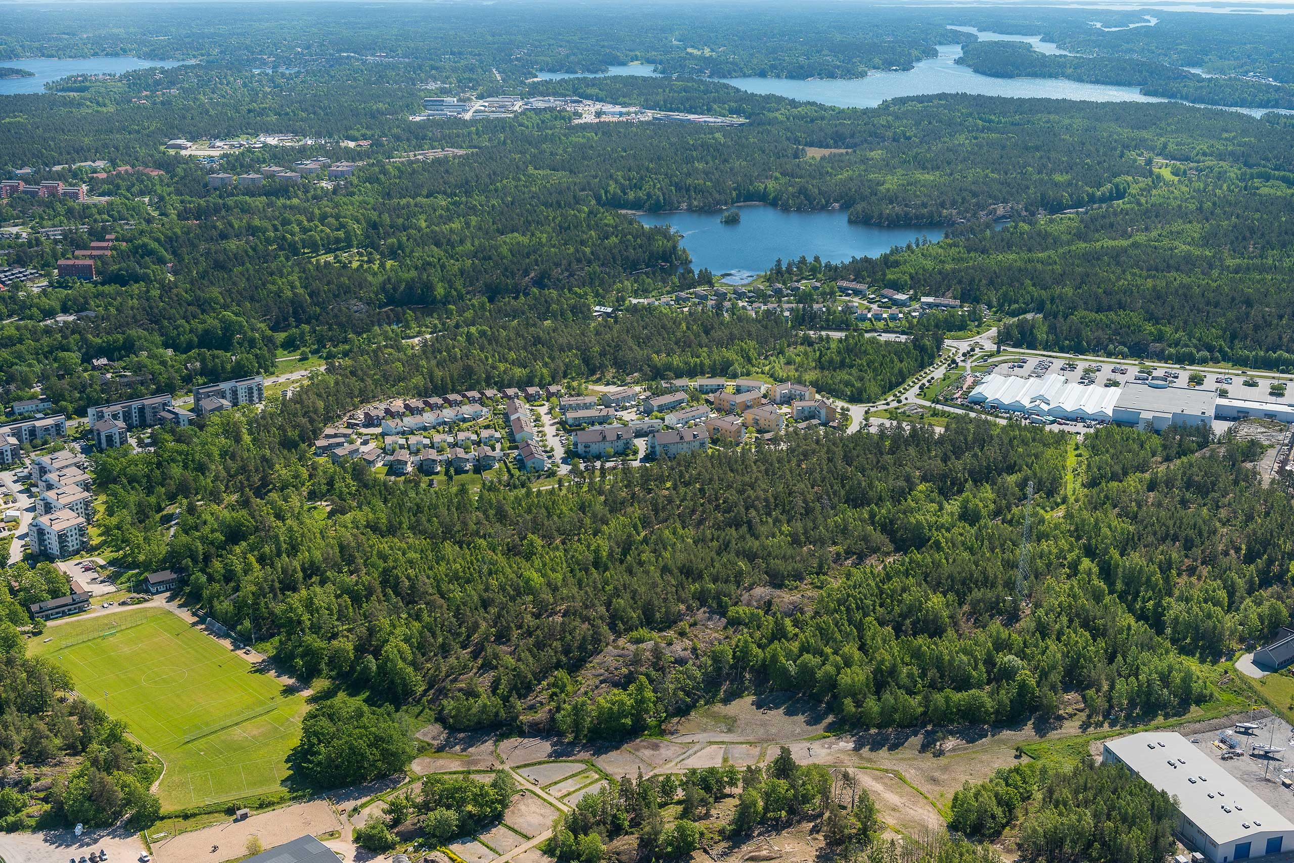 Flygvy över stora skogspartier med insprängda villaområden, ett flertal sjöar och vattendrag syns också, allt är somrigt och grönt.