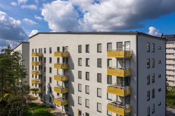 Blå himmel och sol över vitt flerbostadshus med gula balkonger.